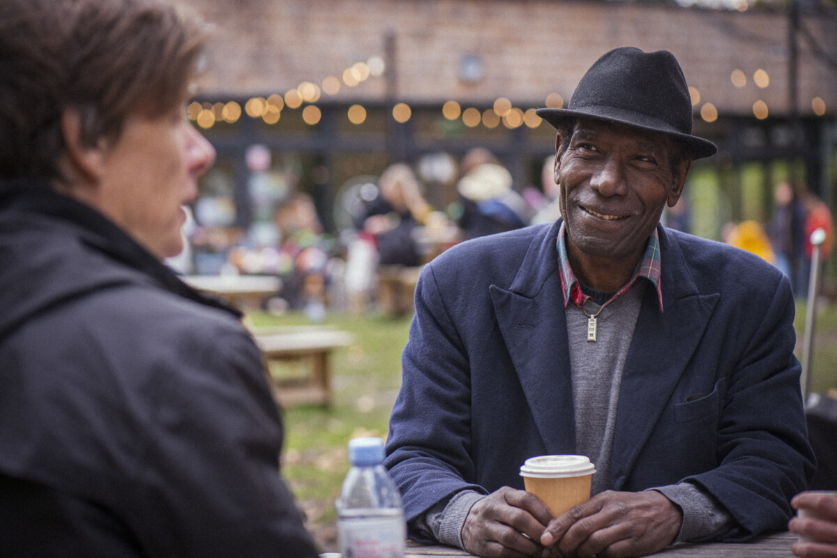Older man with hat, smiling drinking a coffee outdoors at an event. Photo: Peter Kindersley. Source: Centre for Ageing Better