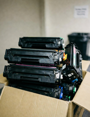 A pile of used printer cartridges in a cardboard box await recycling in an office. Created with Grok AI.