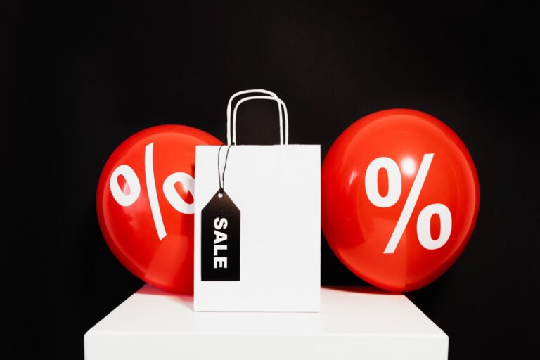 Shopping bag with two red balloons with percentage symbol, against a black background. A black sale label hangs off the paper bag's handle.