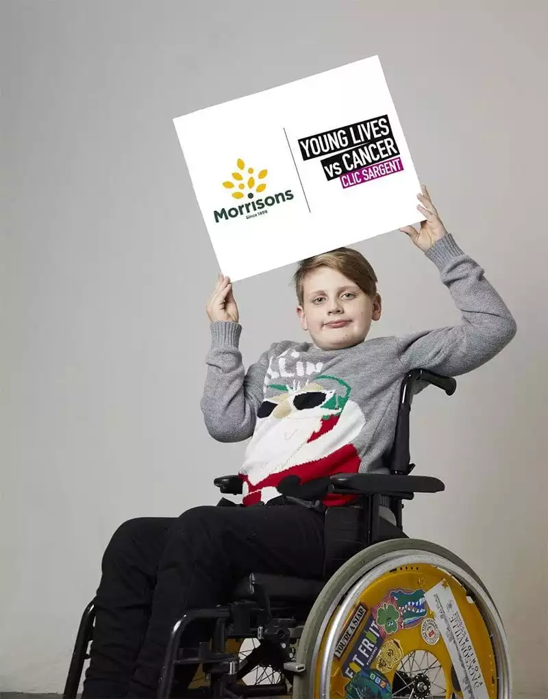 Noah Bailey-Moloney in wheelchair holding Morrisons / CLIC Sargent partnership board