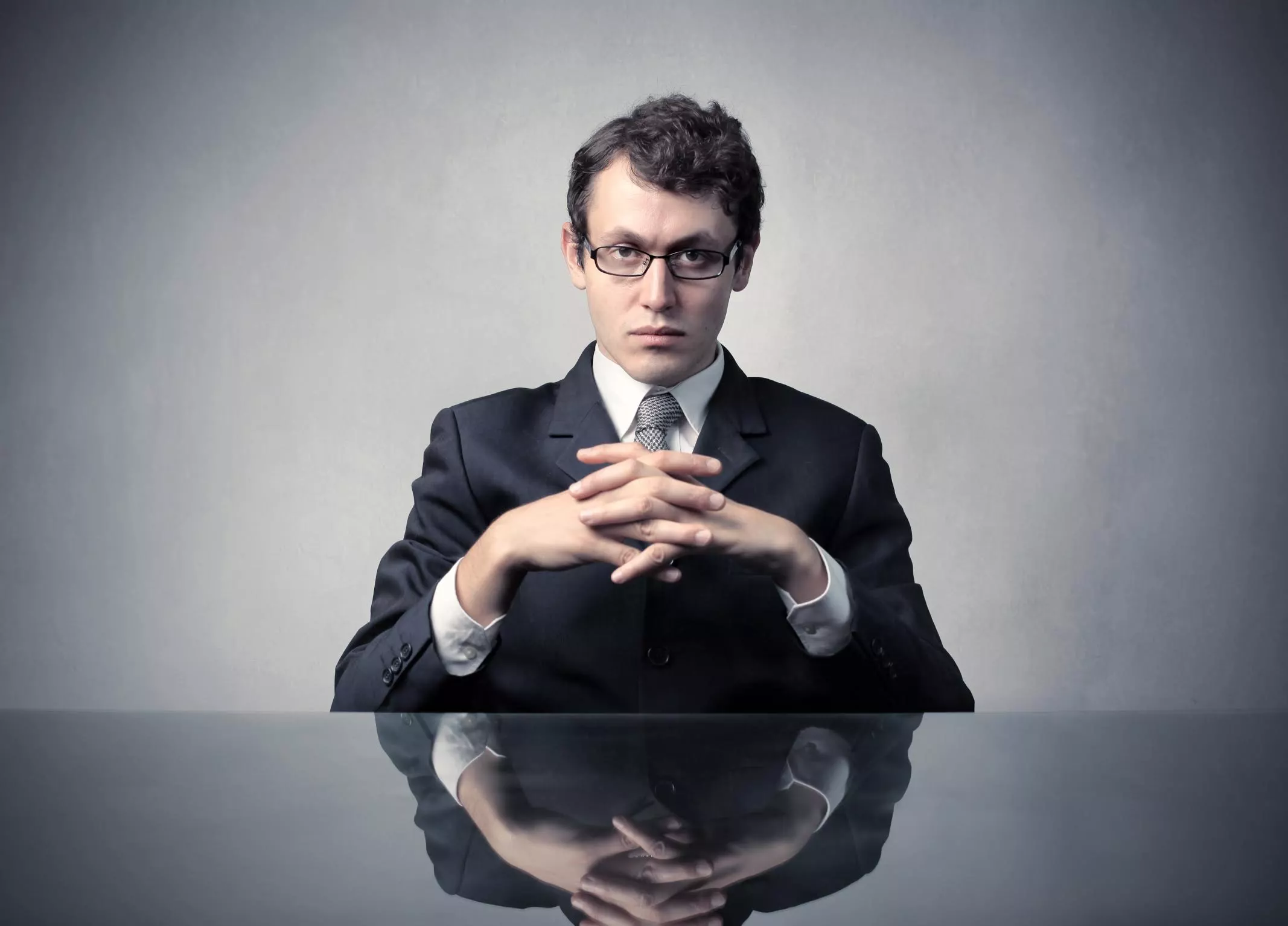 Serious-faced man sitting at table, clasping hands. Image: Shutterstock via Giles Pegram