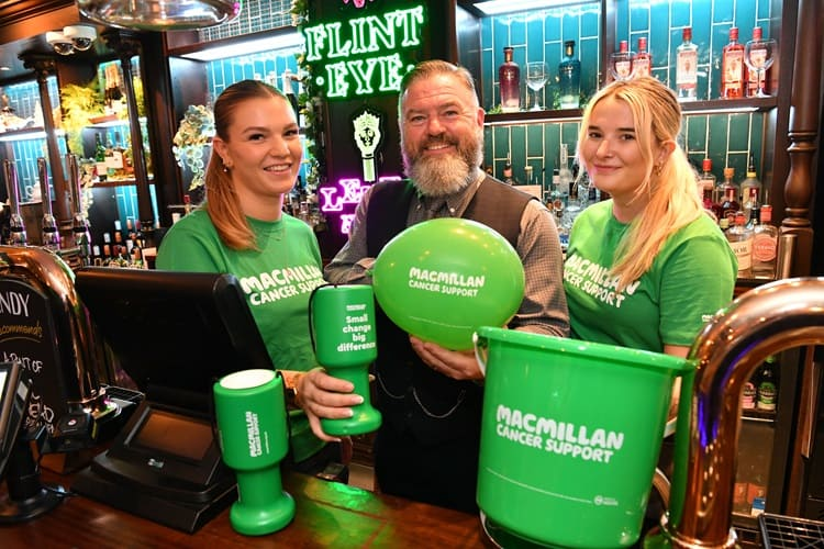 Greene King team members fundraising for Macmillan Cancer Support. Three staff members behind the bar holding green Macmillan collecting tins and buckets.