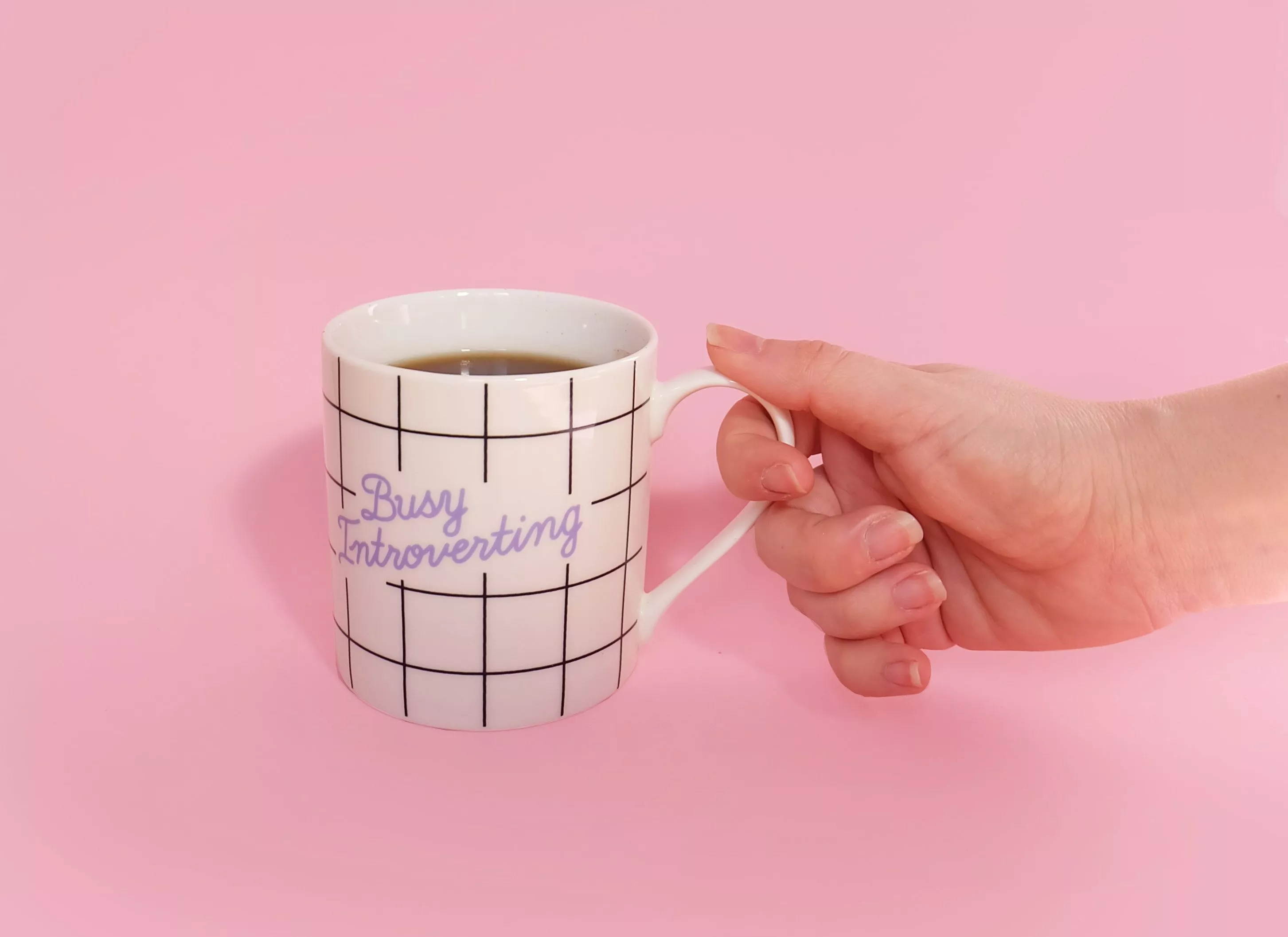 Mug with "busy introverting" on the outside, held by one hand, on a pink background. Photo: Unsplash