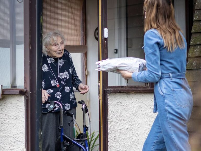 Woman in blue denim jumpsuit delivering food to woman with walking frame standing at her door