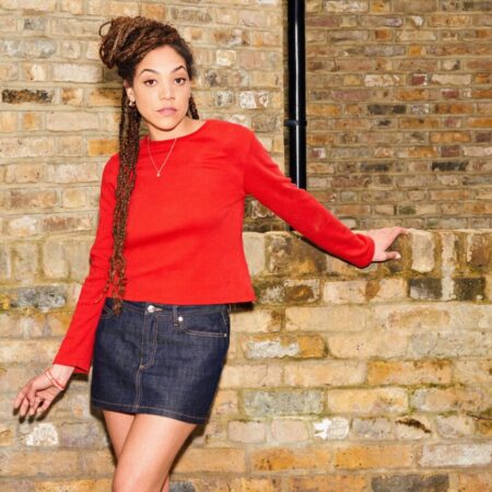 Miquita Oliver in a red jumper & denim mini skirt against a brick wall