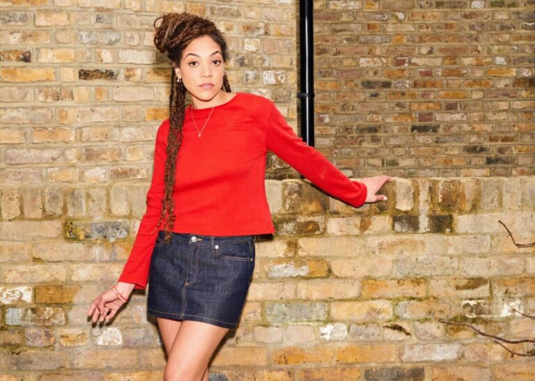 Miquita Oliver in a red jumper & denim mini skirt against a brick wall
