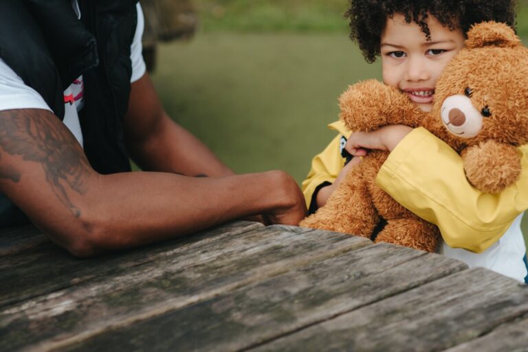 toddler with his dad sitting at a picnic table, hugging a teddy bear. Photo by Anete Lusina from Pexels