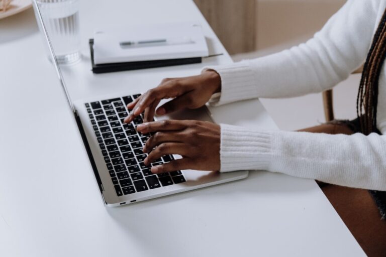A black woman in a white long-sleeved top sits at a desk typing on a laptop - photo: Pexels