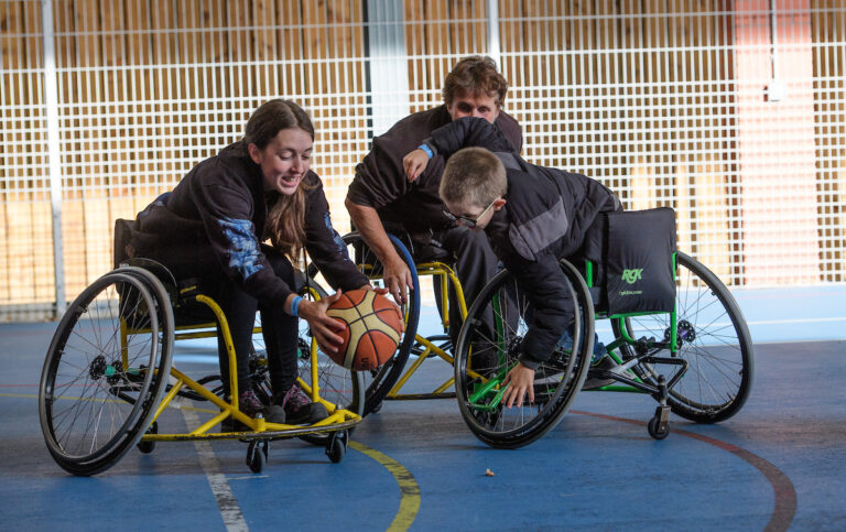 Two young adults and one boy in wheelchairs play basketball on a court