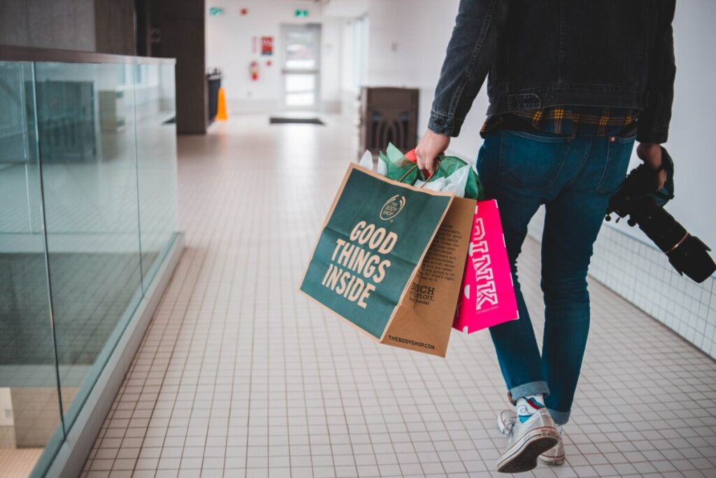 'Good things inside' on the outside of one of two shopping bags held by a man carrying a camera and walking beside a glass panel in a shopping centre - photo: Unsplash