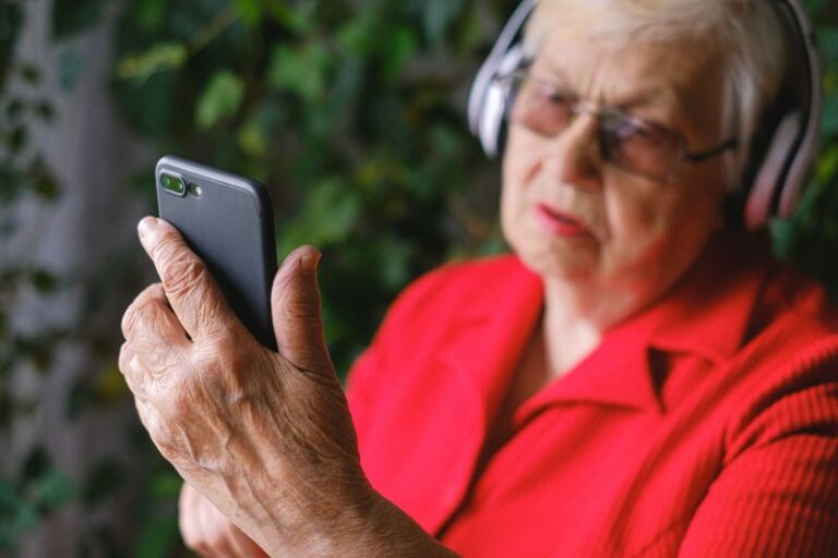 An elderly lady in a red top and glasses, wearing headphones, looks at her smartphone