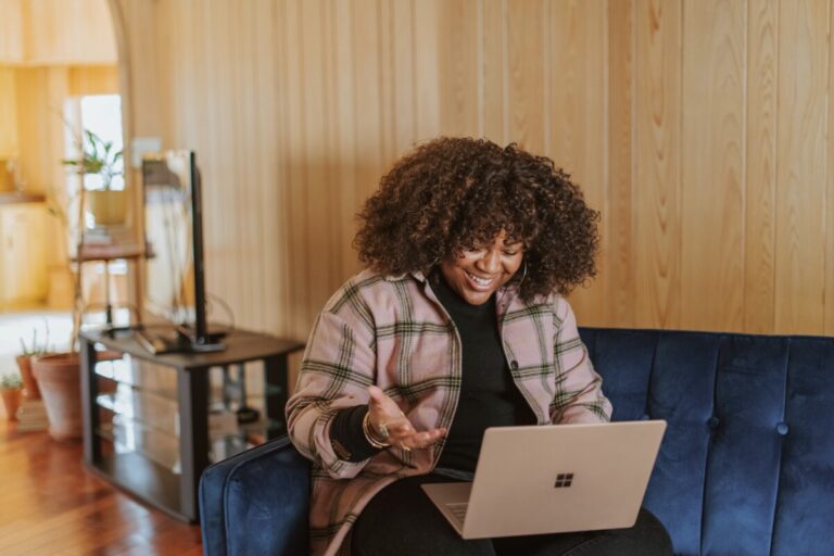 A Black woman with shoulder length hair and a checked shirt sits on a blue sofa, with her laptop, smiling, on a video call