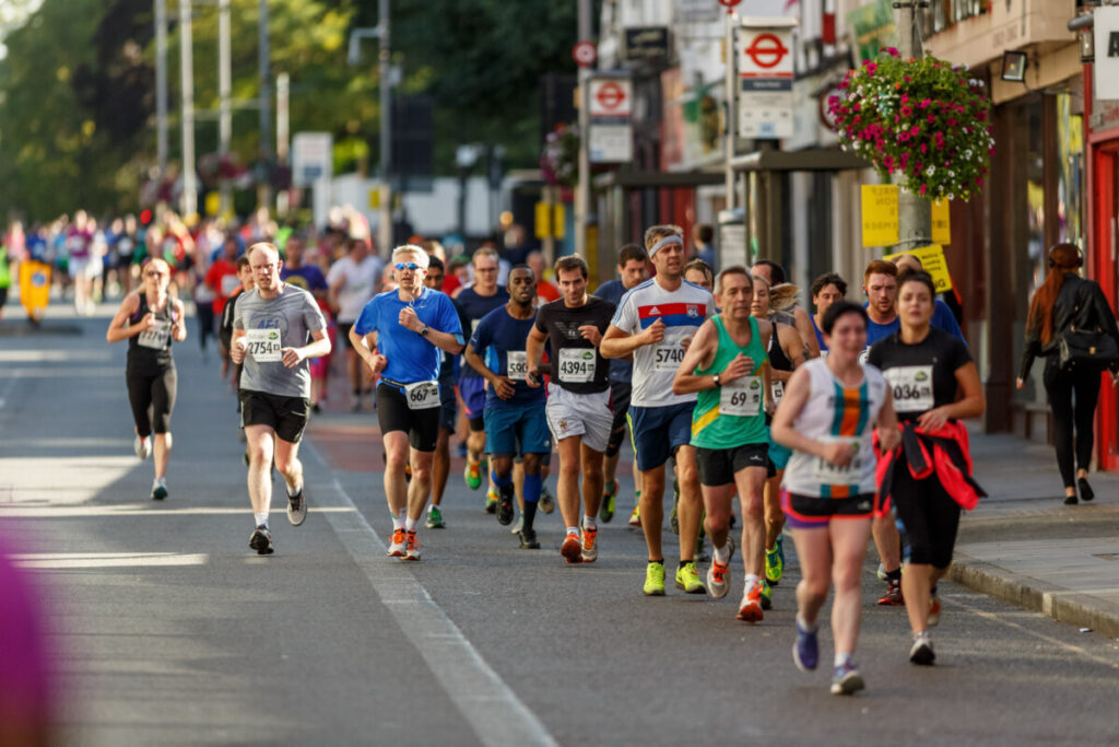 Runners in the Ealing Half Marathon, on a sunny day, running downhill past shops and bus stops. Photo: George Blonsky