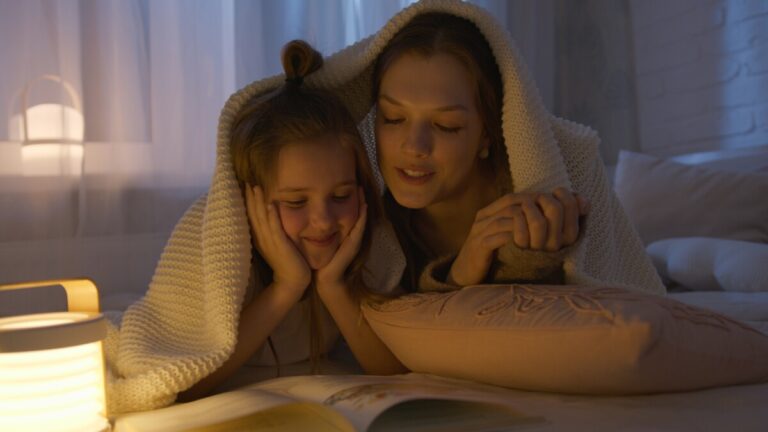 A woman and child reading under a blanket, with the gentle light of a lamp