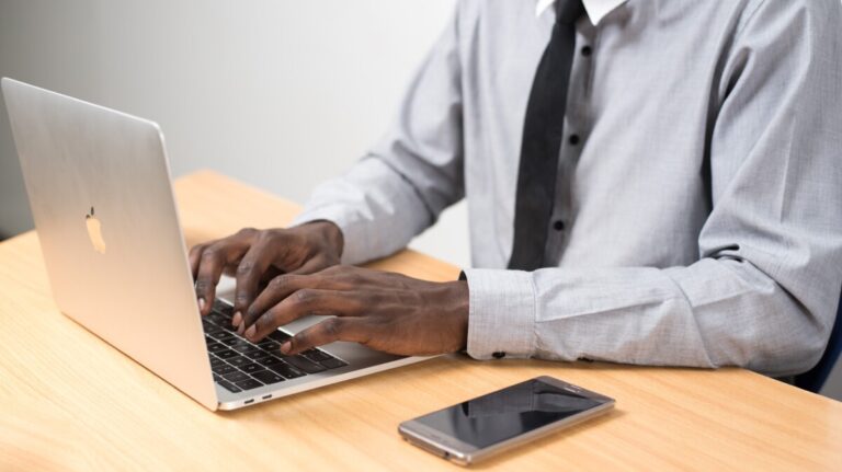 A man in grey shirt and black tie types on his laptop on a wooden desk
