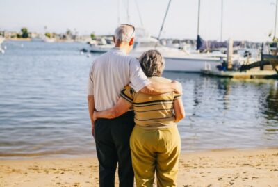 An older couple wrap their arms around each other and gaze at a boat from the sandy beach