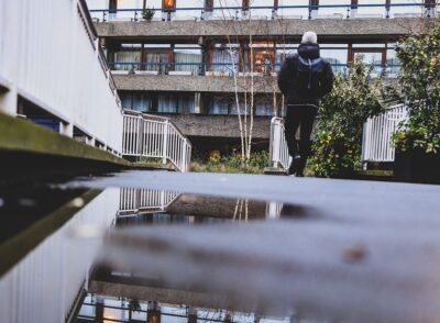 a man walks towards a block of flats, which is reflected in a puddle