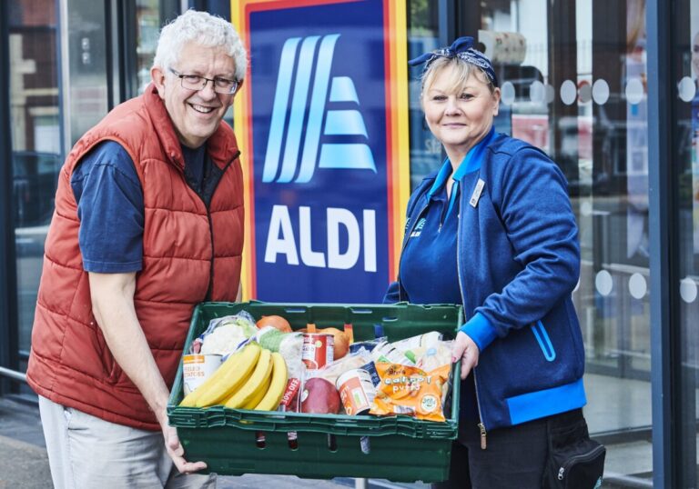 An aldi member of staff hands a crate of fruit and vegetables to a man in front of a blue Aldi sign