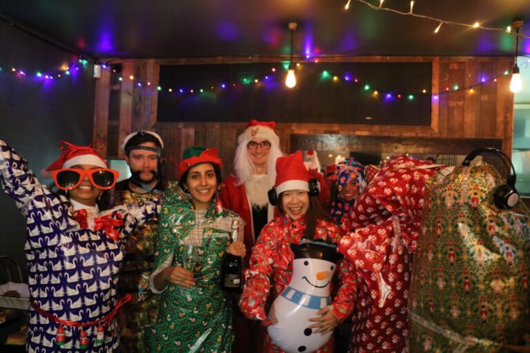 A group of people in Santa hats and dressed in christmas wrapping paper smile at the camera,