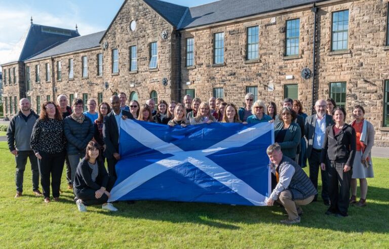 A group of people from Foundation Scotland stand on the grass holding a Scottish flag outside a building