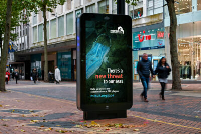 A digital advertising screen on a high street shows an ad from the Marine Conservation Society illustrated by a disposable mask on the beach