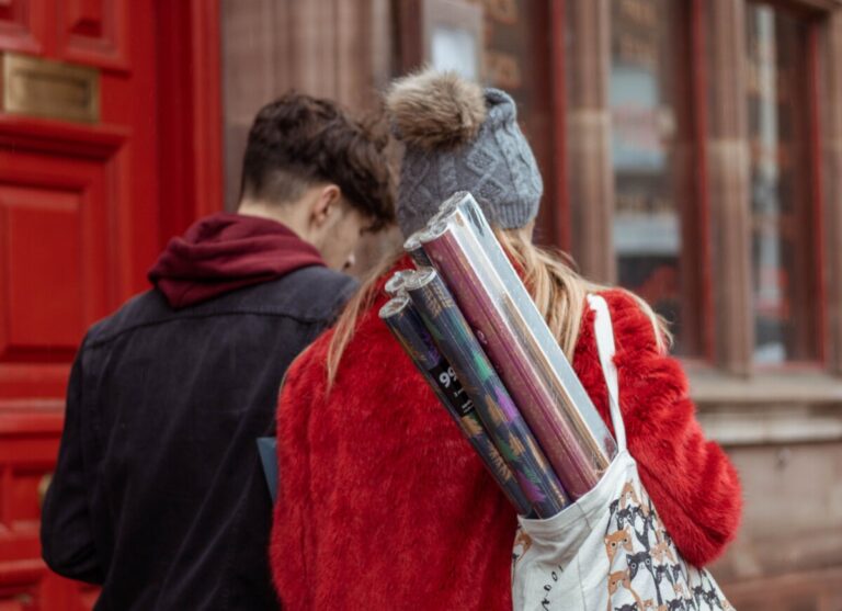 A young couple walk away from the camera, past a red door. The young woman is carrying a bag over her shoulder filled with rolls of christmas wrapping paper