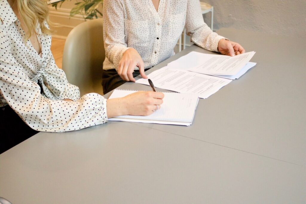 two women both in white shirts sit at a table, one taking notes on a notepad, and the other leafing through documents