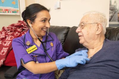 A nurse in a purple uniform listens to the heart of an elderly man, both sitting on a sofa