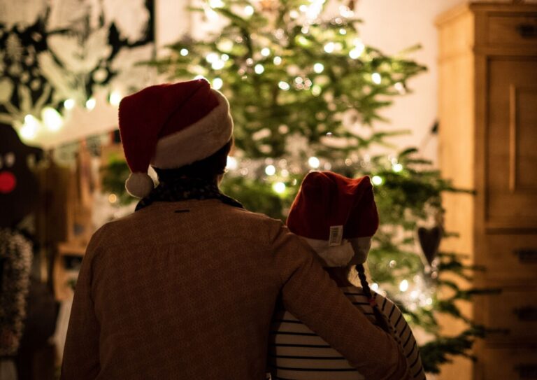 A parent and child in santa hats sit in front of a christmas tree with its lights glowing