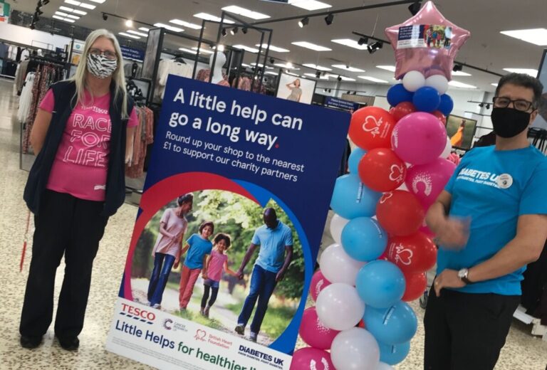 Two tesco employees stand by a sign promoting a fundraising campaign for BHF, Cancer Research UK and Diabetes UK