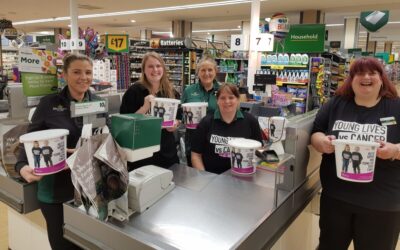 A group of Morrisons colleagues stand by a till, wearing Young Lives vs Cancer t-shirts & holding donation collection buckets