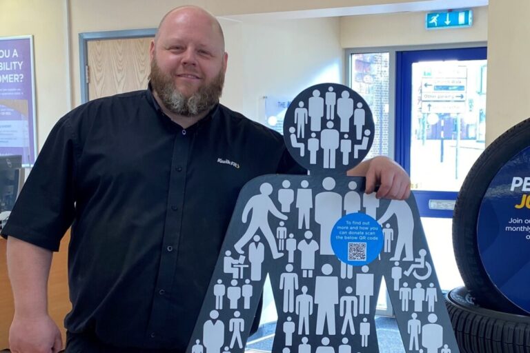 A man with a beard wearing a Kwik Fit polo shirt stands with his arm around a Prostate Cancer UK cut out of a man - its man of men