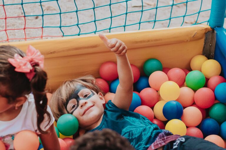 A young boy smiles and holds his thumb up in a ball pit. He has a black eye mask painted on his face