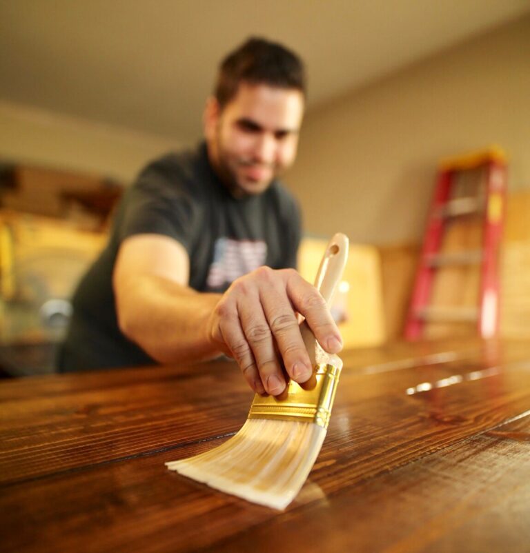Man varnishing a table with a brush, with some step ladders resting against a wall behind him. Photo: Pexels.com