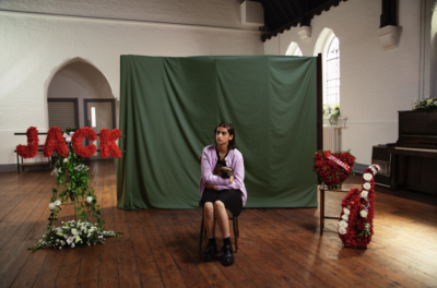 A young girl sits in front of a screen, by red funeral flowers for a man called Jack, in a hall