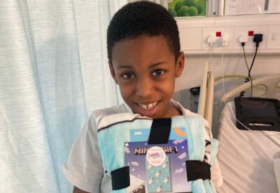 A young boy smiles in front of his hospital bed as he holds a minecraft blanket