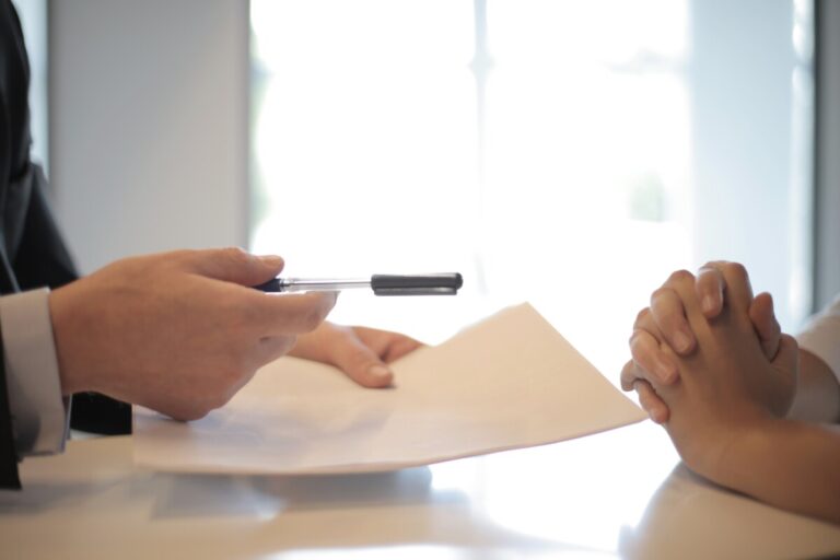 A man's hand holds a sheet of paper while his other hand offers a pen to a clasped pair of hands