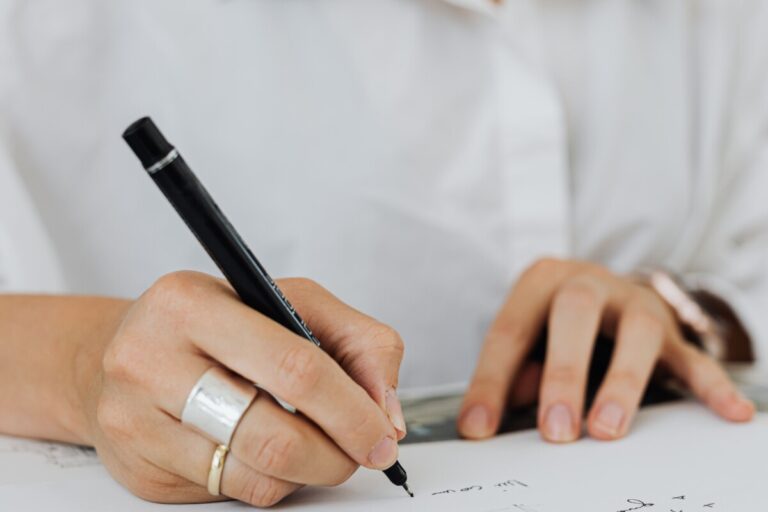A woman wearing silver rings and a white shirt writes with a black pen