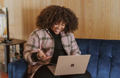 A woman with shoulder length curly hair and a pink checked shirt sits on a blue sofa with her laptop on her lap, smiling at the screen during a video call