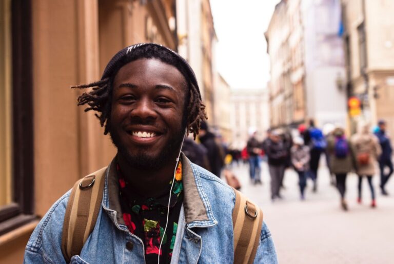 A Black male student smiles at the camera as he walks down a city street with people in the background
