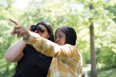 Two young Black women stand in a wood, as one directs her binoculars where the other one is pointing. From Children Nature Network on Nappy