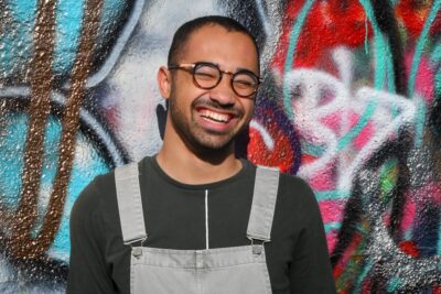 A Black man with short hair and glasses smiles at the camera in front of a wall painted with street art. By Max Harlynking on Unsplash