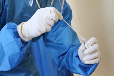 A blue-gowned healthcare worker holds a covid testing swab