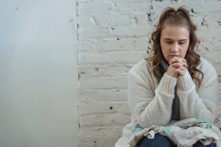 A young woman with long brown hair sits with her hands clasped, looking worried, against a white brick wall. Photo by Keira Burton from Pexels