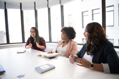 Three business women talk around a table in a meeting room. Photo by Christina Morillo on Pexels