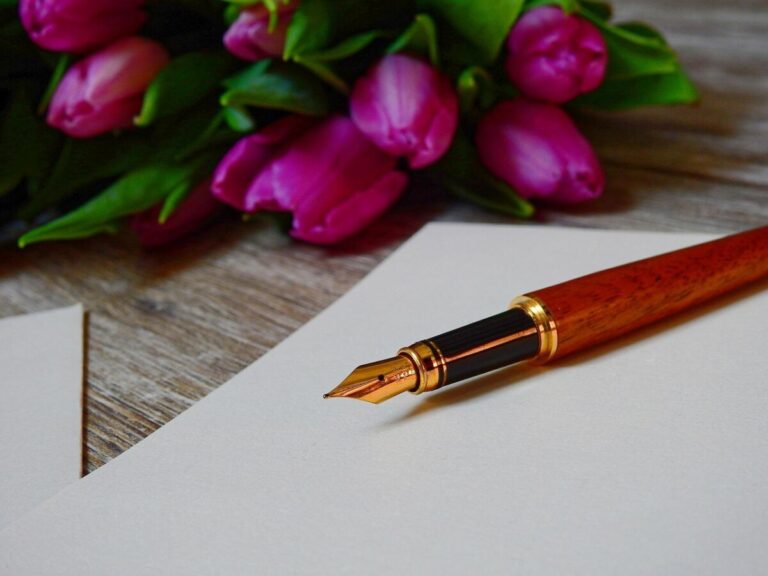 a fountain pen rests on a sheet of paper, in front of a bunch of pink tulips