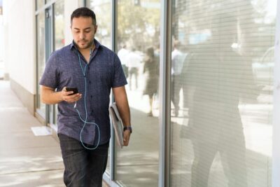 Man in tshirt walking down a sunny street, listening to a podcast on his phone. Image: Pexels.com