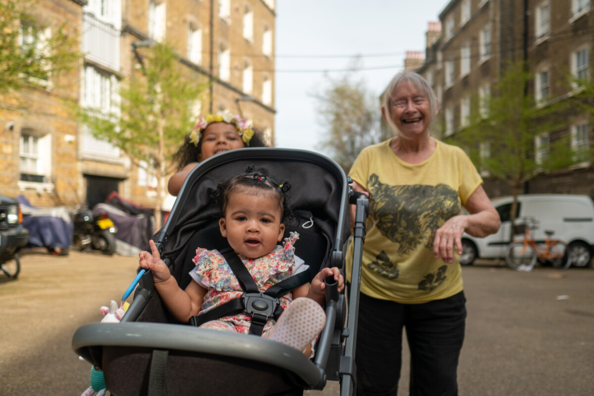 Older woman smiling with grandchildren, one in a pushchair, outside between housing blocks. Photo: Elliot Manches, Centre for Ageing Better