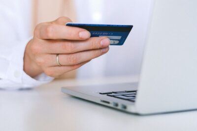A woman's hand holds a debit card in front of a laptop. By Anna Shvets on Pexels