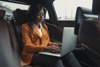 Woman wearing an orange blazer with a laptop open sitting in a car. Photo: Tima Miroshnichenko at Pexels.com